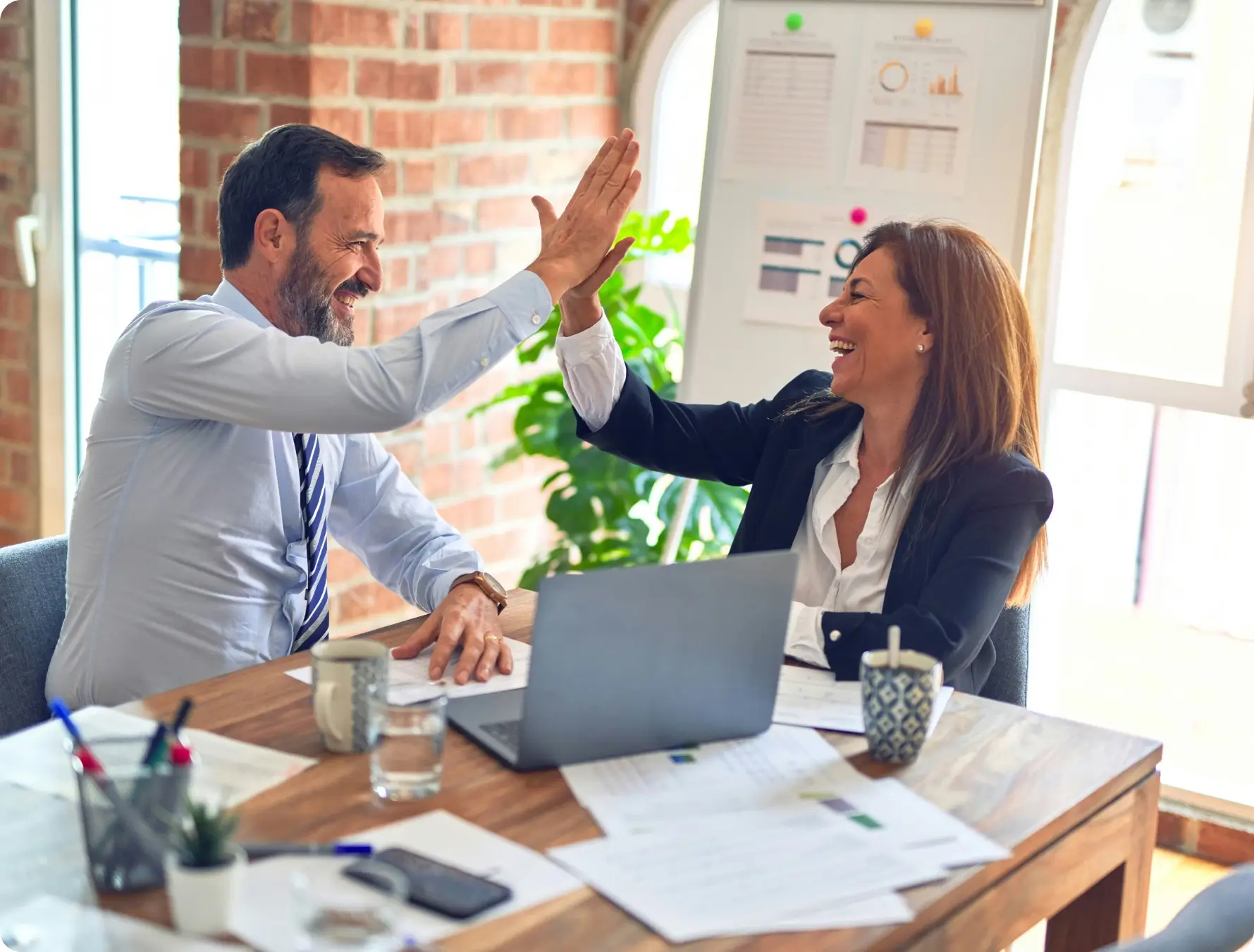 Two business professionals, a man and a woman, smiling and giving each other a high five over a desk with papers, a laptop, mugs, and charts in a bright office setting.