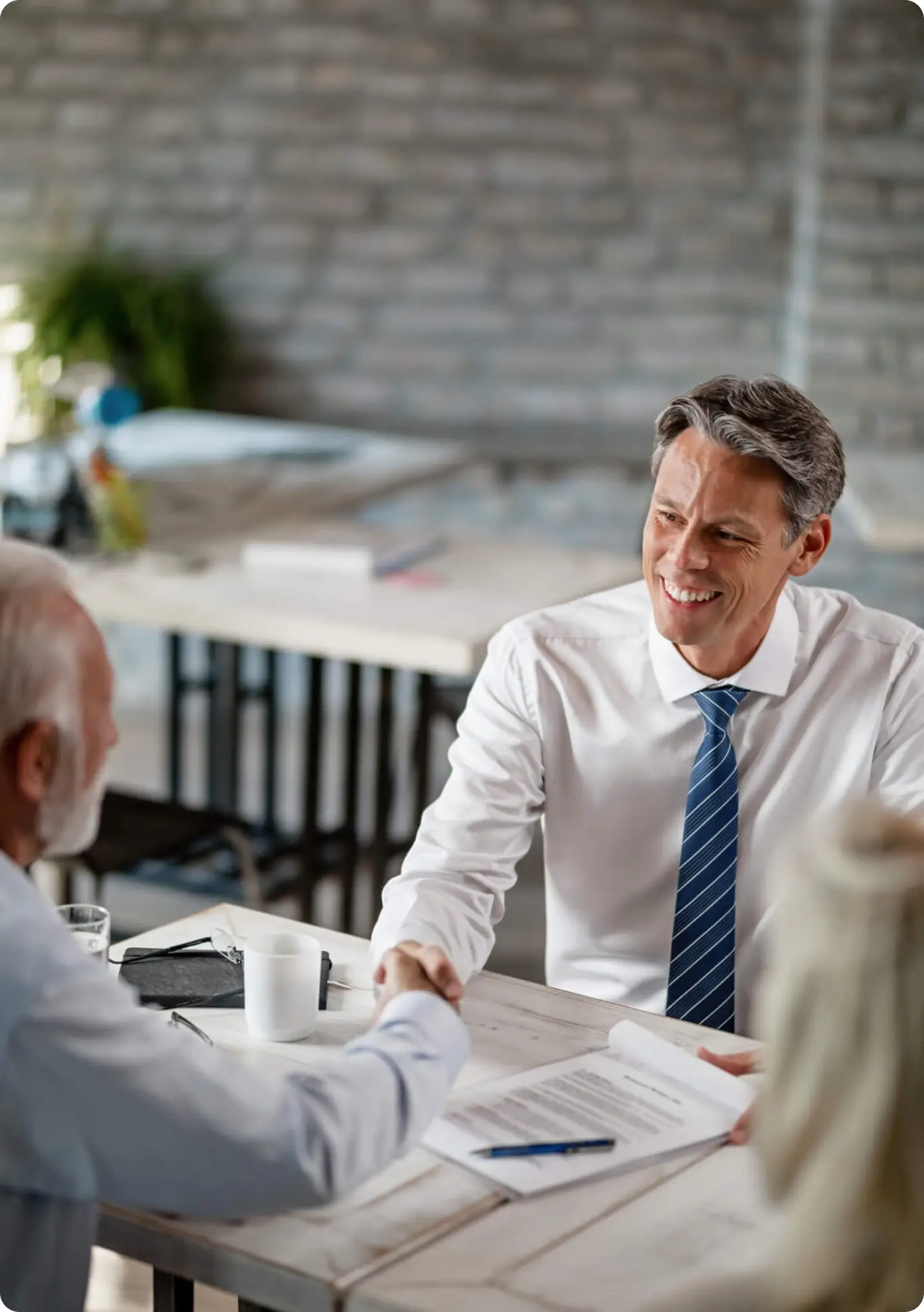 Two men in business attire sit at a table shaking hands and smiling, with documents and a pen on the table, suggesting a successful business meeting discussing financial strategy solutions in a modern office setting.