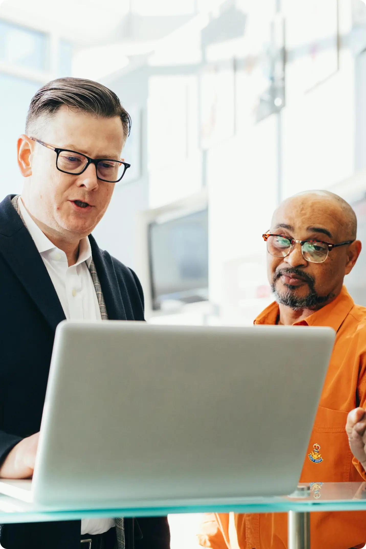 Two men standing in front of a laptop, engaged in discussion. One man wears a dark jacket and glasses, while the other wears glasses and an orange shirt. Bright indoor setting with blurred background.