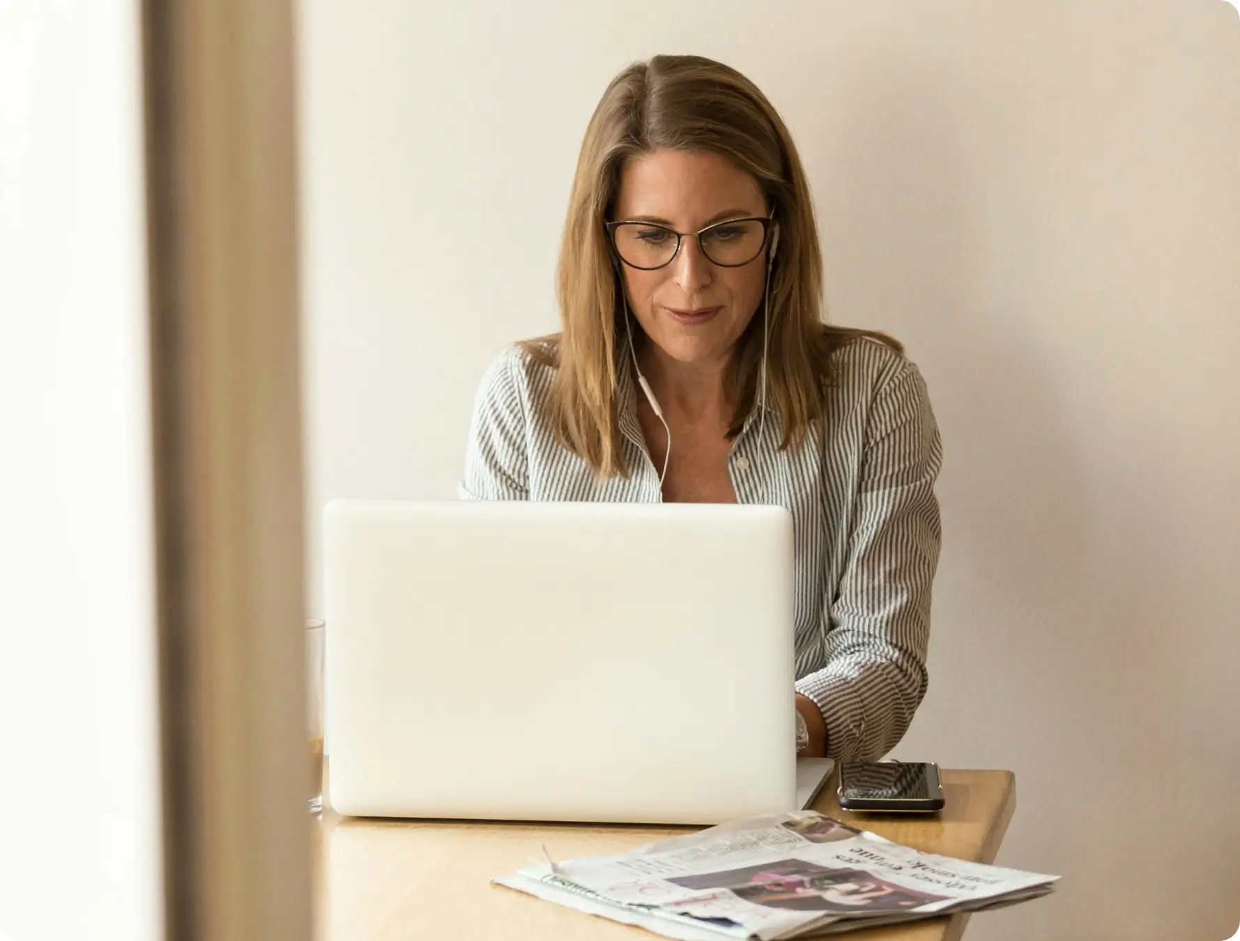 A woman with glasses and a striped shirt works on a laptop at a wooden table, with newspapers and a smartphone beside her, in a well-lit room with a light-colored wall.