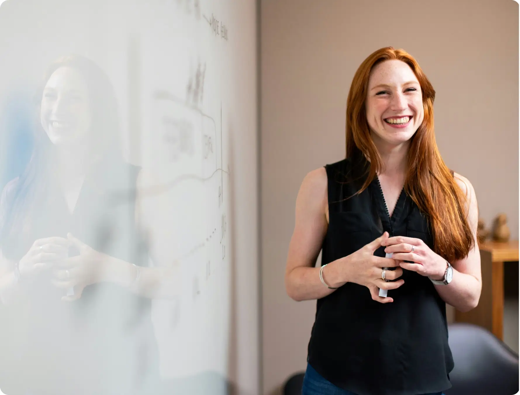 A woman with long red hair, wearing a black sleeveless top, stands smiling in an office next to a whiteboard with faint writing and diagrams, her hands clasped in front of her.